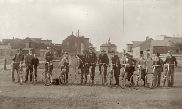 Thirteen men and boys pose in a line with their bicycles. Most of the group poses with similar bicycles, except for one man standing with a penny-farthing or ordinary bicycle. In the background are houses, and a large brick building with a clock tower.