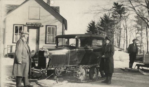 Winter scene with Chester Thordarson (left) and Bill Jepson standing next to a vehicle with skis attached to the front, and three crawler wheels in the back. The Washington Island Post Office is behind them.
