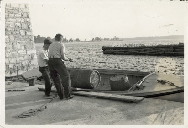 Two men unload barrels from a boat at a boathouse.