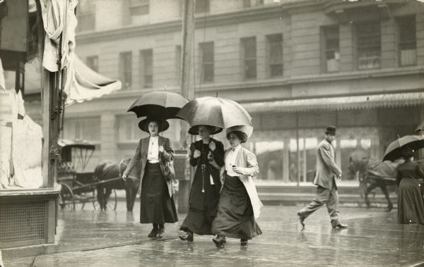 Three women in hats and long skirts walk with umbrellas in the rain.