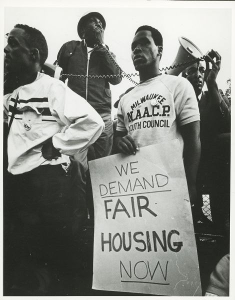 Protesters at a rally for fair housing. One man is holding a sign that reads: "We Demand Fair Housing Now," while others behind him are using a bullhorn.