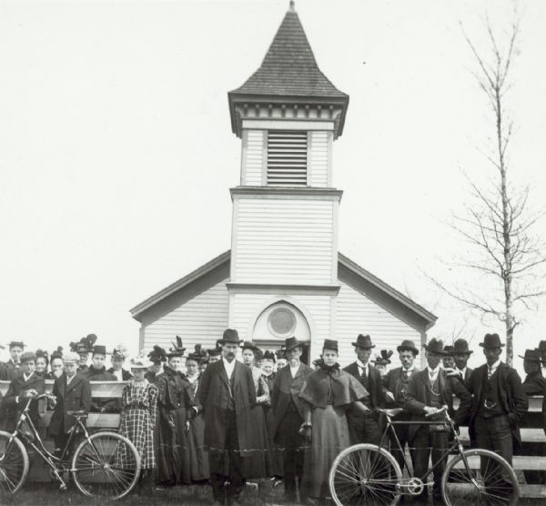 A group of well-dressed people posing with bicycles in front of the Waupun Norwegian Lutheran Church, also called the Norske Evangelical-Lutherske Kirke, which was built in 1855  This is probably the congregation of this church.