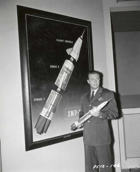 William Lundigan, who plays Colonel Ed McCauley on the television show <i>Men Into Space</i>, holds a model of a flight capsule and rocket. A large poster of the rocket and capsule is on the wall next to him. He is dressed in a military uniform.