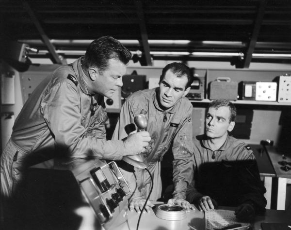 William Lundigan, as Colonel Ed McCauley on the television show <i>Men Into Space</i>, talks into a microphone. Two other men are next to him, one standing and the other sitting. All three men wear flight suits.