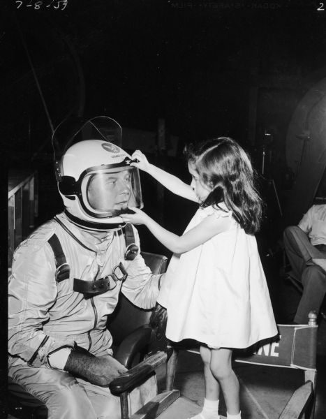 William Lundigan is seen with his daughter on the set of the television show <i>Men Into Space</i>. He is wearing a spacesuit and helmet. His daughter is trying to lift the visor of the helmet.
