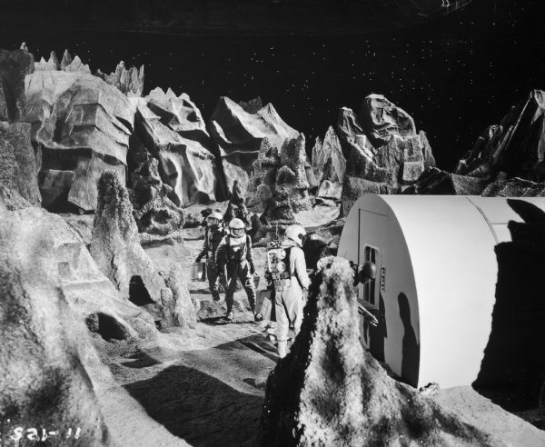 Four actors, dressed as astronauts, are seen on the set of the television show <i>Men Into Space</i>. The set depicts the lunar landscape and the men are near a building.