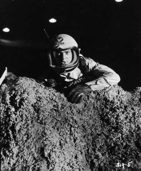 Douglas Dick, as Harold Carter on the television show <i>Men Into Space</i>, peers over a mound of rocks. He is wearing a spacesuit and helmet.