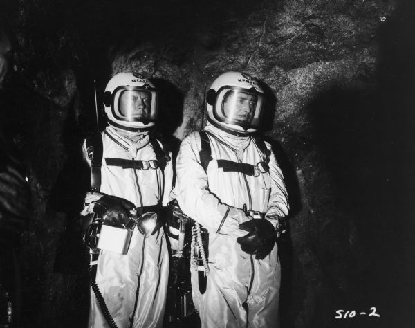 William Lundigan, as Colonel Ed McCauley, and John Howard as Dr. Rowland Kennedy, both dressed in space suits, stand next to each other in what appears to be a cave.