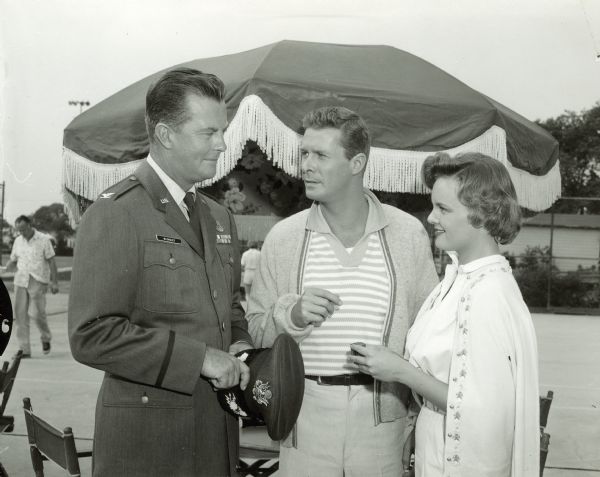 William Lundigan, as Colonel Ed McCauley, stands next to Fred Beir, who played Lt. Art Frey, and Nan Peterson, who appeared as Ann Hammond on an episode of the television show <i>Men Into Space</i>. They are outside on a tennis court and Lundigan is wearing a military uniform.