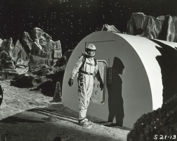 William Lundigan, as Colonel Ed McCauley on the television show <i>Men Into Space</i>, stands outside a building on the lunar landscape set of the show. He is dressed in a spacesuit and helmet.