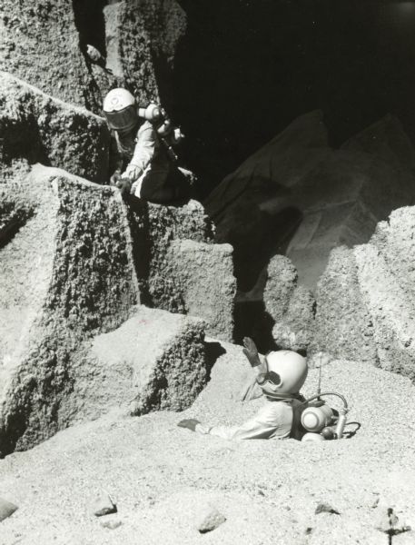 Two actors dressed as astronauts are seen on the lunar landscape set of the television show <i>Men Into Space</i>. One is perched on some rocks while the other is buried up to his chest in dirt.