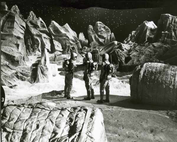 Three men dressed in astronaut suits stand on the lunar landscape set of the television show <i>Men Into Space</i>. A spaceship can be seen in the background.