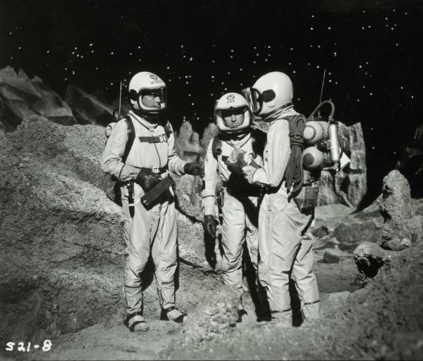 William Lundigan as Colonel Ed McCauley, Don Ross as Dr. Orr, and James Coburn as Dr. Narry, stand on the lunar landscape set of the television show <i>Men Into Space</i>. They are all wearing spacesuits and helmets.