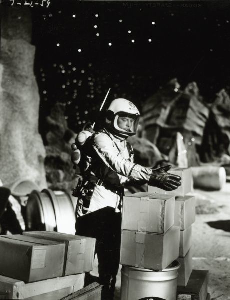 William Lundigan, as Colonel Ed McCauley on the television show <i>Men Into Space</i>, stacks some boxes. He is wearing a spacesuit and helmet and appears to be on the lunar landscape set of the show.