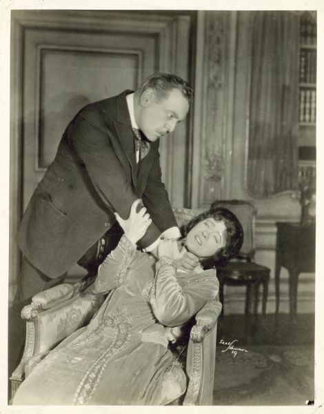 Ruth Chatterton and Henry Miller are shown in a scene from the play <i>La Tendresse</i>. Chatterton sits in a chair while Miller stands above her with his hands around her neck.