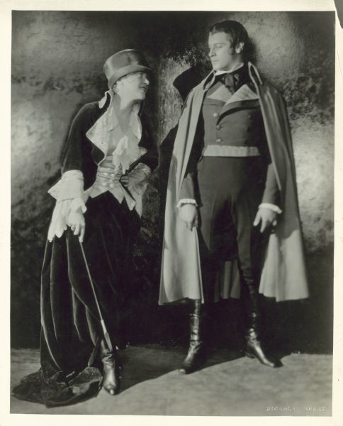 Ruth Chatterton and Ralph Forbes posing in costumes from the play <i>The Man With a Load of Mischief</i>. Chatterton holds a riding crop and wears a long velvet coat, a frilly long-sleeved blouse, a top hat and boots. She smiles and looks up to Forbes who is wearing a cape over a suit.