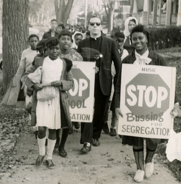 Father James Groppi marches with African American teenage girls who are carrying signs that read, "Stop Bussing For Segregation" and "Stop School Segregation".