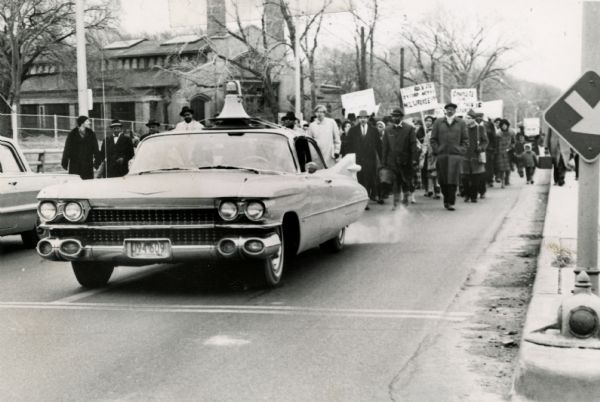 A group of protesters march down a street, led by a car. They are protesting school segregation.