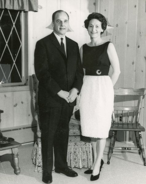 Full-length portrait of Governor Gaylord Nelson and Mrs. Carrie Lee Nelson. They are standing in front of an upholstered chair and a wooden chair in a wood paneled room.