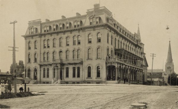 An exterior view of the Park Hotel, at the corner of Main and Carroll Streets. Children and adults are standing on the street corner on the left. A large group of people is standing on the balcony and porch of the hotel. Further down the street is the large steeple of a church.