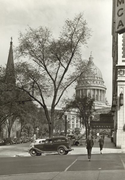 View down sidewalk along West Washington Avenue, looking toward the Capitol. A man is crossing the street and there is a car at the corner that has the words "Read The Capitol Times" on the side. On the next block is an awning for the Loraine Hotel. On the extreme right side, part of a YMCA sign is on the building on the corner.