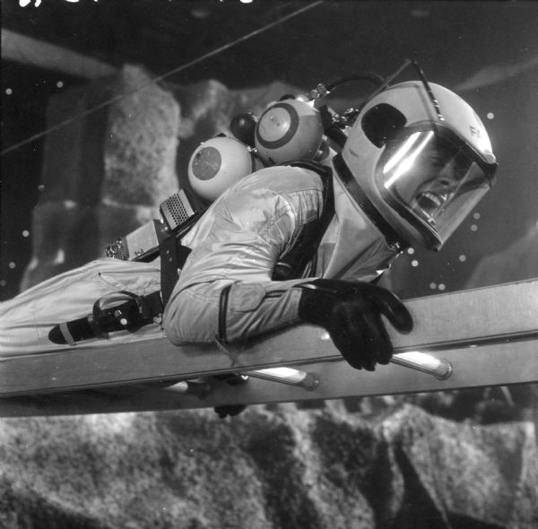 Arthur Franz, as Captain Tom Farrow on the television show <i>Men Into Space</i>, lays on top of a ladder. He has a look of pain on his face and is dressed in a spacesuit and helmet.