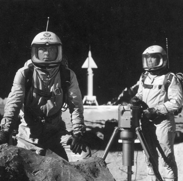 William Lundigan, as Colonel Ed McCauley, and Ross Elliott as Dr. George Batton, stand on the lunar landscape set of the television show Men Into Space. They are both dressed in spacesuits and helmets. A spaceship can be seen in the background.