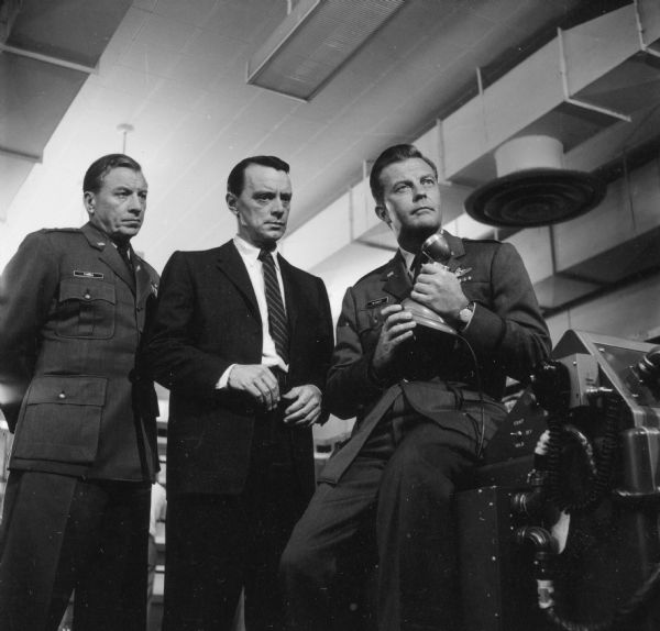 Ken Lynch, as General Thomas, Harry Townes, as Dr. William Thyssen, and William Lundigan as Colonel Ed McCauley, are seen on the television show <i>Men Into Space</i>. Lundigan holds a microphone and sits on a desk.
