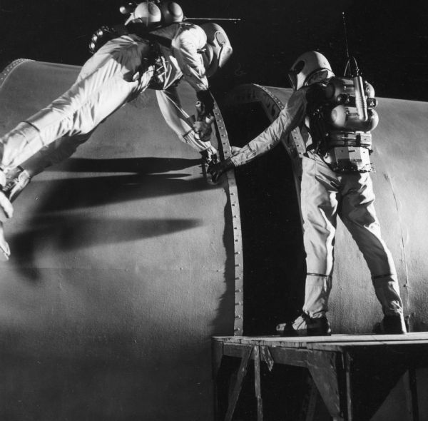 Two actors, dressed as astronauts, are seen on the set of the television show <i>Men Into Space</i>. The two look like they are trying to repair something. One stands on a platform and the other is suspended next to him.