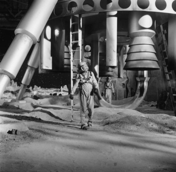 An actor, dressed as an astronaut, is seen on the lunar landscape set of the television show <i>Men Into Space</i>. He is standing near the leg of a huge spaceship. Another actor can be seen in the background underneath the ship.