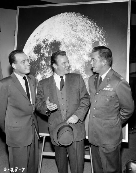 William Lundigan, who plays Colonel Ed McCauley on the television show <i>Men Into Space</i>, is seen talking to two other men. He is wearing a military uniform, and the three men stand in front of a large photograph of the moon.