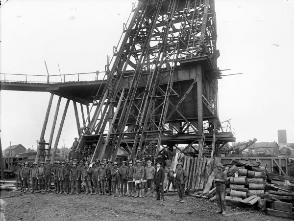 Group portrait of workers posing outside a mine, probably in Hurley, Wisconsin. Three young boys stand behind the group among stacks of logs.