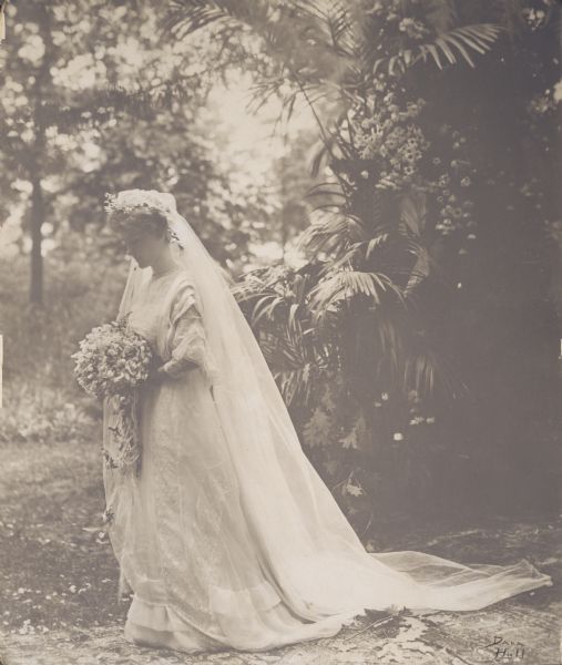 Portrait of Josephine Crane of Chicago, Illinois in her wedding dress and veil. She married Harold Cornelius Bradley, a professor of physiological chemistry at the University of Wisconsin, in Lake Geneva, Wisconsin on July 8, 1908. She is standing outdoors on what appear to be carpets.