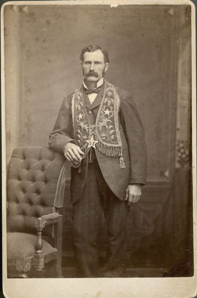 Three-quarter length studio portrait of Harlow Waller standing and wearing a star-shaped medal and and embroidered ceremonial collar over his suit. He is probably a member of the Independent Order of Odd Fellows.