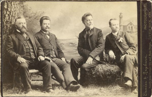 Studio portrait of four men sitting in front of a painted backdrop. From the left are Vojta Masek and Charles Jonas sitting on a bench. The men sitting on the hay bale on the right are unidentified.