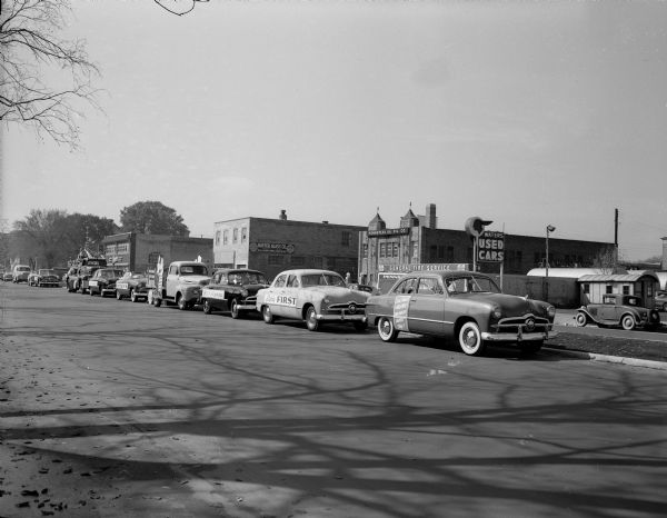 Line-up of Ford cars and trucks for the Kayser safety parade in the 700 block of East Washington Avenue. The banner on the lead car reads "Enter Ford's $100,000 CAR SAFETY CONTEST". During this contest any automobile, regardless of make, can be brought to any Ford dealer for a free safety check.