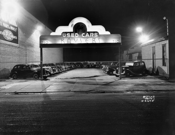 Kayser's R&G Used Car lot at night with neon sign on arch at 710 E. Washington Avenue. There is also a sign painted on the side of a brick building on the left.