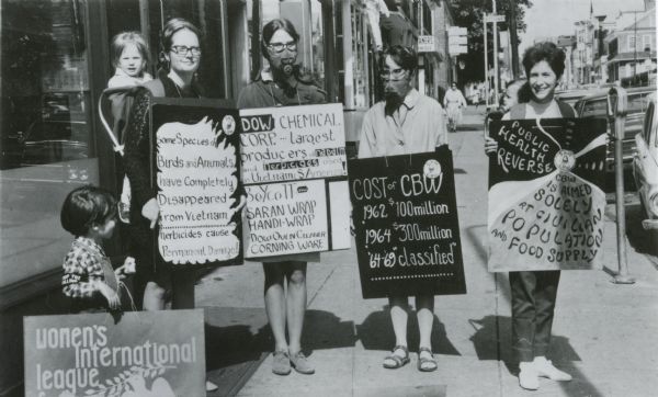 A small group of women, along with two children, hold signs protesting Dow Chemical. Two women are wearing gas masks.