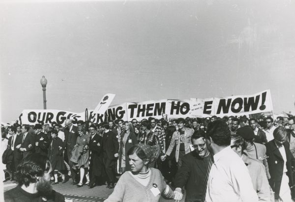 A crowd of people gather to demonstrate. They hold a banner that reads, "Our GI's Bring Them Home Now!".