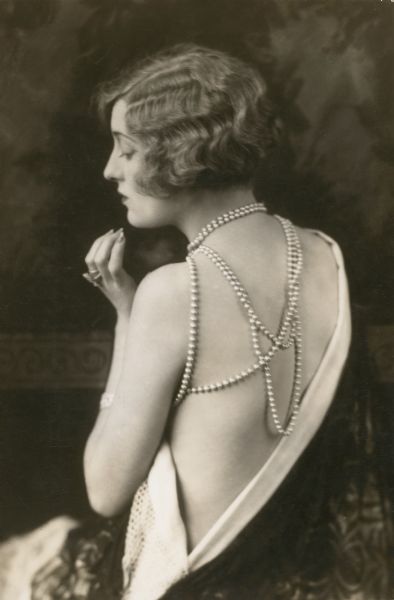 Waist-up portrait of actress Peggy Hopkins Joyce. Her back is to the camera and her face is in profile. She wears a one-shoulder dress that exposes most of her back. She wears a strand of pearls around her neck and shoulder that drapes over her exposed back.