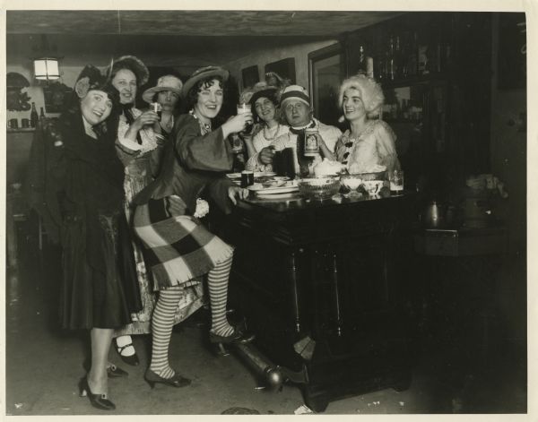 Seven people in costumes posed around a bar and holding glasses of beer.