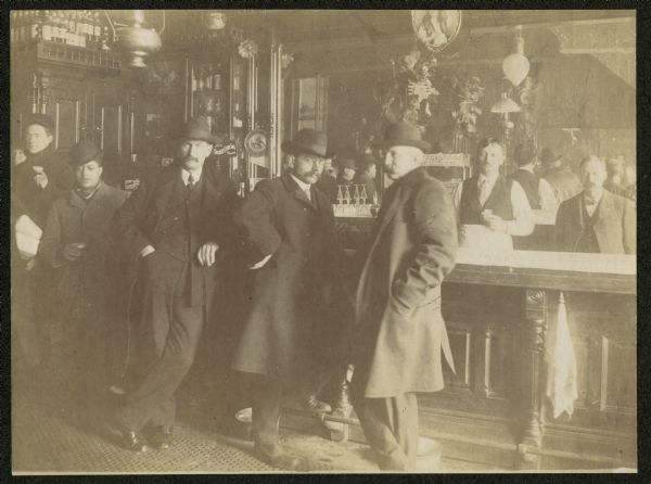 Group of men posed at a bar, 3 Star Saloon located at 224 Main Street. The bar owner Azard Bunker is standing to the far right behind the bar. The bartender is standing behind the bar, which has a large mirror behind the cash register.