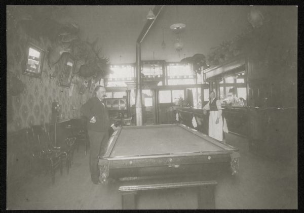 Man standing with his hand on a pool table in the center of a bar with a high ceiling. The bartender leans against the bar on the right. The room is decorated along the ceiling and walls with numerous taxidermied animals and framed pictures. In the background is a woodstove, and the entrance with a door framed with stained glass windows.