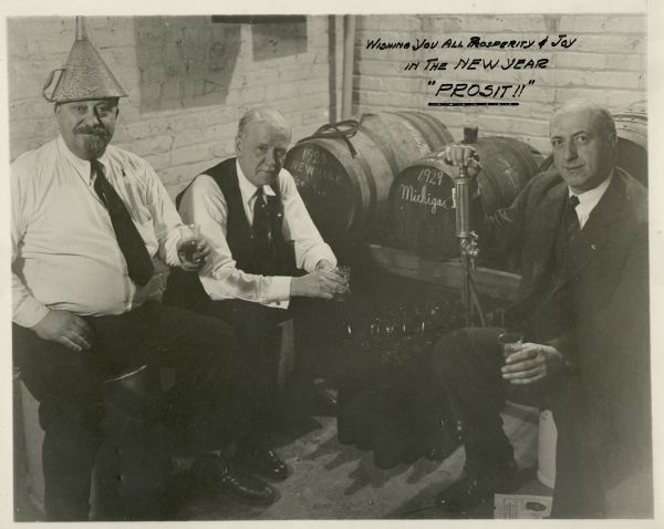 Group portrait of three men seated near beer casks, each holding a small glass. One man wears a funnel as a hat. The photograph is inscribed with the toast, "Wishing you all prosperity and joy in the New Year. 'Prosit!!'" The casks are marked 1929 New York and 1929 Michigan.