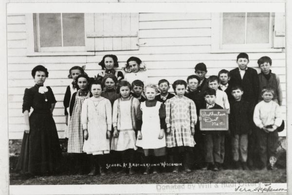 Group of students and a teacher posed outside a school building. One boy holds a chalkboard sign reading "Brothertown Dist. No. 2."