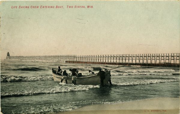 Group of men on a life saving crew launching a rowboat into Lake Michigan. A long pier with a lighthouse at the end bisects the horizon. Caption reads: "Life Saving Crew Entering Boat, Two Rivers, Wis."