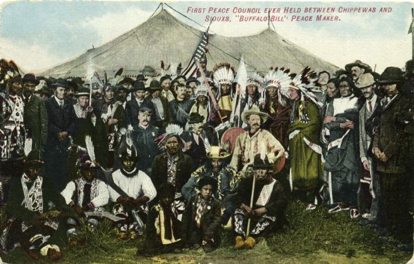 Colored postcard view of "Buffalo Bill" Cody center right in white coat and hat surrounded by Native Americans in traditional garb, military personnel and other onlookers. The group is seated in front of a large tent. A U.S. flag flies behind the group.