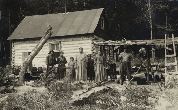 Potawatomi family standing in front of a log cabin. Elders identified the woman in the center as Mrs. Frank Young, and the man on the right as community leader White Pigeon.
