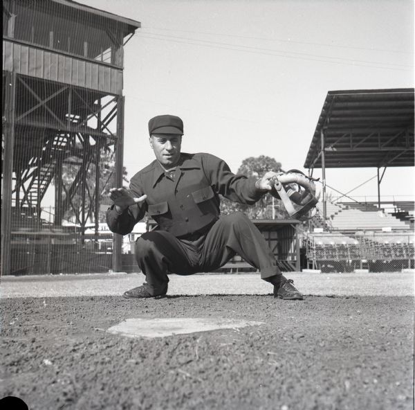 Umpire, Augie Donatelli, posing in a crouch behind home plate and demonstrating the umpire signal for 'safe.' He is holding his protective mask in his hand.