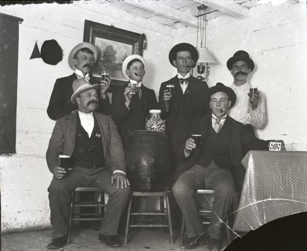 Six men posed around a beer keg, holding glasses of beer. One man has a deck of cards.